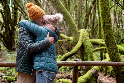 Old senior couple enjoying nature outdoors in a mountain forest with moss covered trunks. Joyful elderly couple in winter clothes embracing in Garajonay national park of La Gomera