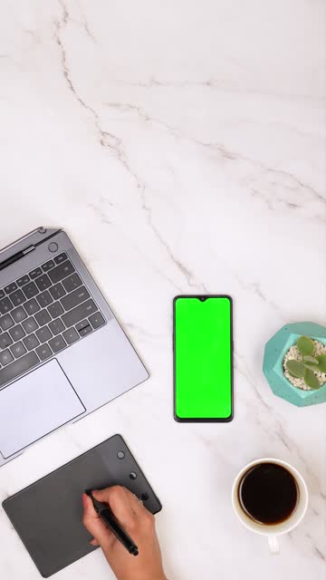 Person watching a video tutorial on mobile phone (green background chroma key) while working with laptop and drawing tablet and drinking coffee. Copy Space above. Vertical video shot top view