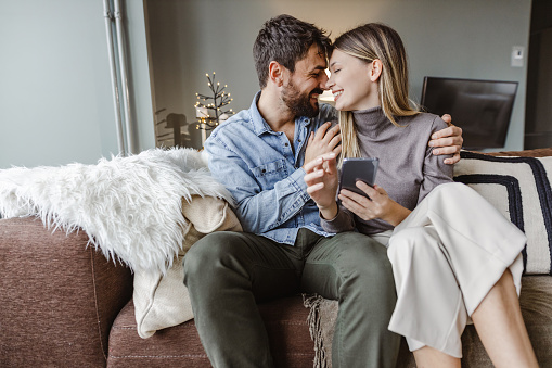 A young couple is in the living room, they are smiling to each other and using mobile phone