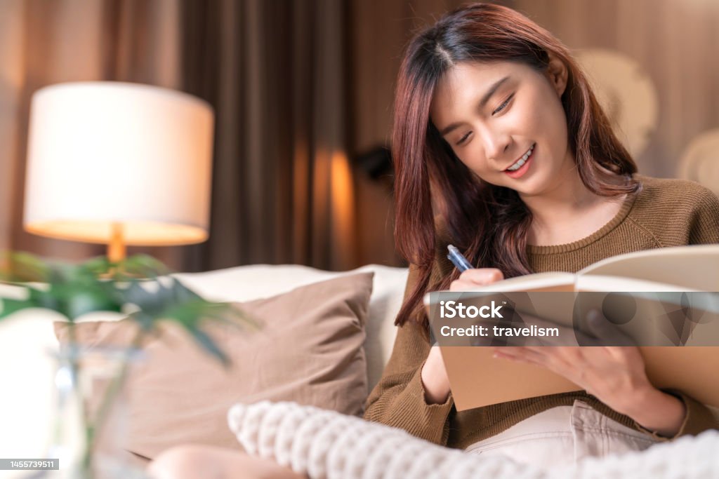 Attractive asian young adult female woman writing in a journal in her home. She diary her ideas and note about her creative work project in living room at home Note Pad Stock Photo