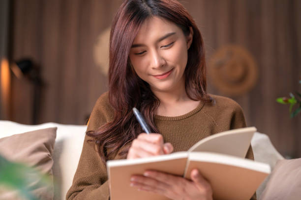 jolie jeune femme adulte asiatique écrivant dans un journal chez elle. elle tient son journal et note son projet de travail créatif dans le salon à la maison - journal intime cocooning photos et images de collection
