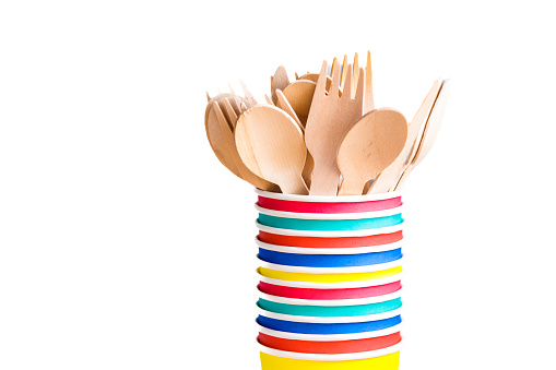 Close up image depicting a collection of recyclable wooden cutlery inside a stack of colorful paper cups on a white background.