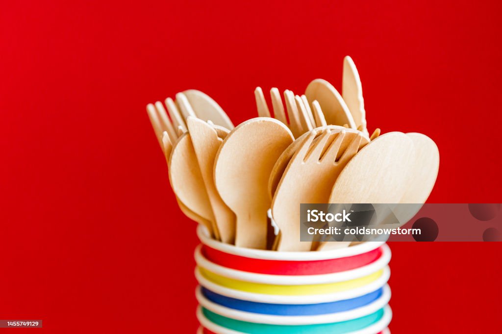 Eco friendly recyclable wooden cutlery and paper cups on red background Close up image depicting a collection of recyclable wooden cutlery inside a stack of colorful paper cups on a red background. Backgrounds Stock Photo