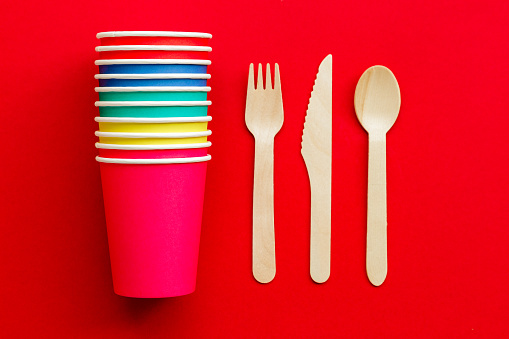Close up image depicting a collection of recyclable wooden cutlery next to a stack of colorful paper cups on a red background.