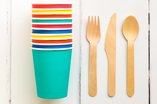 Close up image depicting a collection of recyclable wooden cutlery next to a stack of colorful paper cups on a white wooden surface.