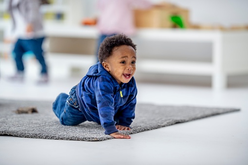 A sweet little boy of African decent is seen crawling around on the floor while at daycare.  He is dressed casually and is smiling as he happily explores his surroundings.