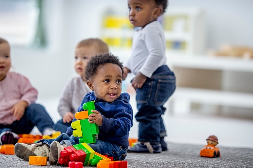 A small group of toddlers sit on the floor of their daycare classroom as they play together.  They are each dressed casually and are playing with colorful blocks.