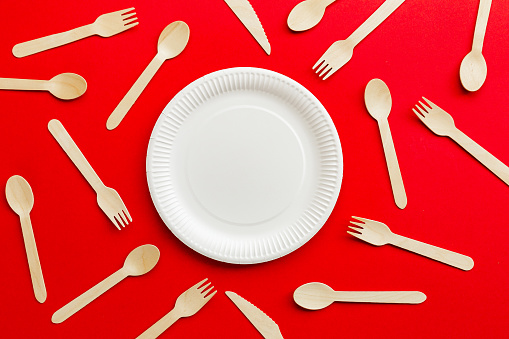 Close up image depicting a collection of recyclable wooden cutlery - knives, forks and spoons - and a paper plate on a red background.