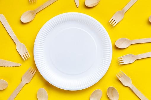 Close up image depicting a collection of recyclable wooden cutlery - knives, forks and spoons - and a paper plate on a yellow background.