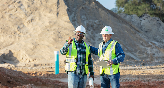Two multiracial men working at a construction site having a meeting. The African-American man, in his 40s, is holding a set of plans. His coworker, a senior man in his 60s, is carrying a digital tablet and walkie-talkie. Mounds of dirt are in the background.