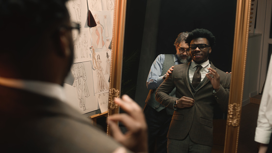African American man on suit fitting in stylish designer atelier or in dim luxury clothing shop. Tailor puts jacket on man in front of the mirror. Tailored wedding or business suit. Fashion concept.