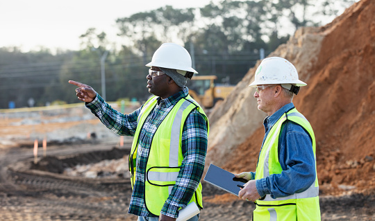 Two multiracial men working at a construction site conversing. The main focus is on the mature African-American man, in his 40s, who is holding a set of plans, pointing his finger. His coworker, a senior man in his 60s, is carrying a digital tablet. A mound of dirt is in the background.