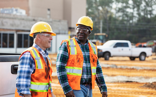 Two multiracial workers walking side by side at a construction site, conversing. The main focus is on the African-American man who is in his 40s. His coworker is a senior man in his 60s.