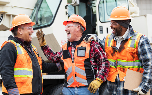 A multiracial group of three construction workers standing in front of a mobile crane, laughing. The one in the middle is a senior man in his 60s. His Hispanic and African-American coworkers are in their 50s. They are all wearing hardhats, protective eyewear, reflective vests and work gloves.