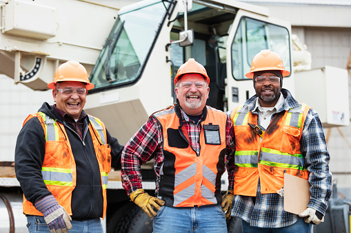 A multiracial group of three construction workers standing in a row in front of a mobile crane, looking at the camera. The one in the middle is a senior man in his 60s. His Hispanic and African-American coworkers are in their 50s. They are all wearing hardhats, protective eyewear, reflective vests and work gloves.