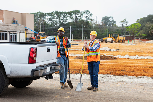 Two multiracial construction workers holding shovels, leaning on pickup truck. The African-American man is in his 40s. His coworker is a senior man in his 60s. They are looking at the camera.