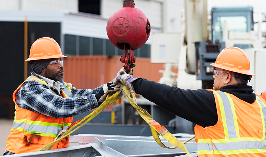 Two multiracial construction workers working together to prepare a large object to be hoisted with a mobile crane. They are attaching the lifting straps to a hook. The man on the left is African-American and his coworker is Hispanic. They are in their 50s.
