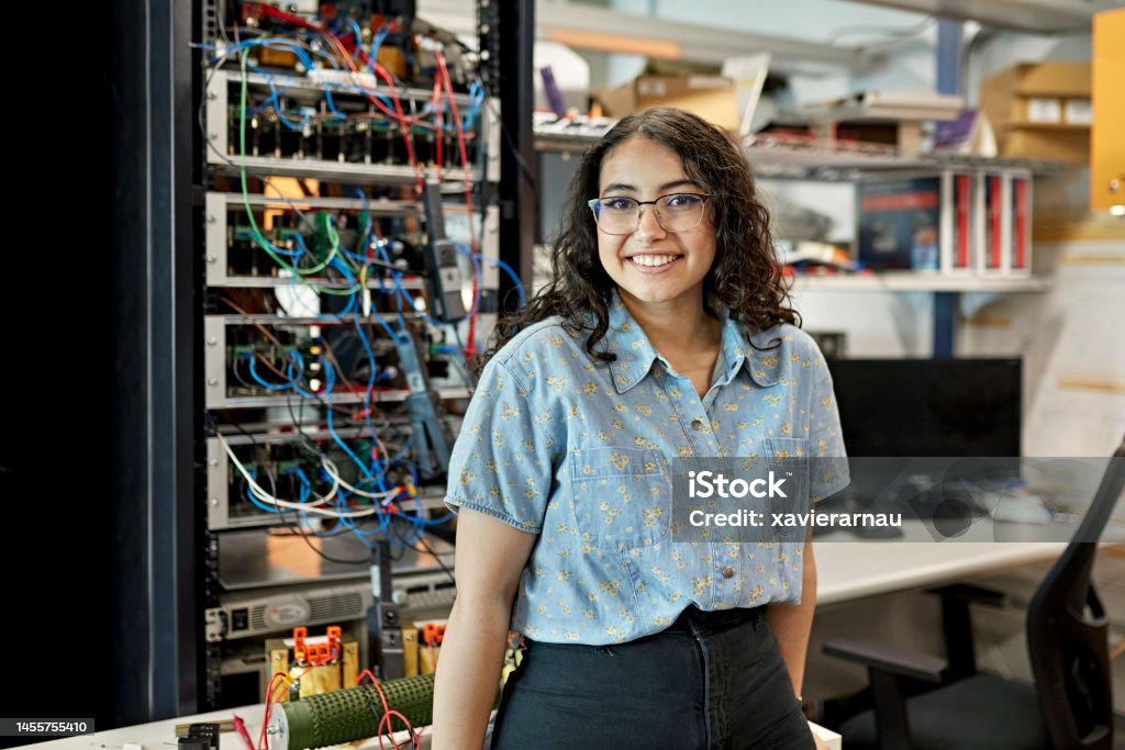 Portrait of early 20s female STEM student Waist-up view of casually dressed woman standing in electrical engineering lab and smiling at camera. Property release attached. Technology Stock Photo