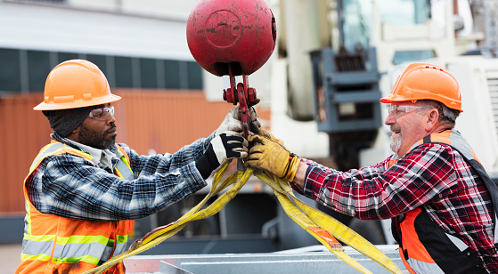 Two multiracial construction workers working together to prepare a large object to be hoisted with a mobile crane. They are attaching the lifting straps to a hook. The African-American man on the left is in his 50s. His coworker is a senior man in his 60s.