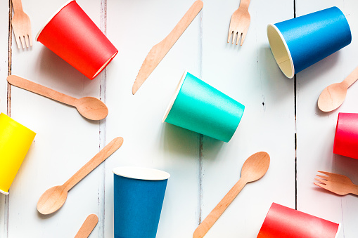 Close up image depicting a collection of colorful and recyclable eco friendly paper cups and wooden cutlery on a white background.