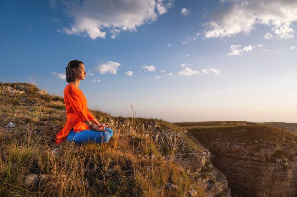A young slender woman in a lotus position in profile in nature in the mountains sits on the grass, meditates, balances near a rock stock photo