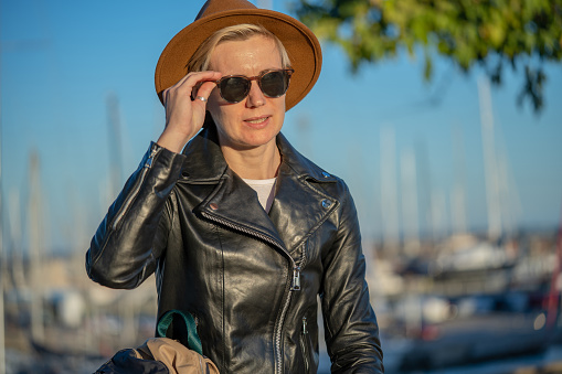 30s blond woman with short hair in classy hat, sunglasses and leather jacket next to yacht marine in Barcelona