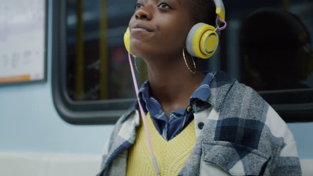 Portrait of Young Black Woman Putting Headphones on in Public Transport and Smiling at Night. Female African American Teenager Using Music Online Platform on Smartphone to Listen to her Favorite Song