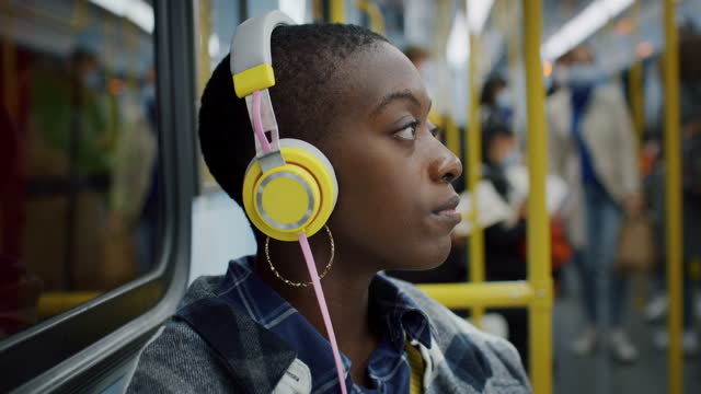 Portrait of Young Black Woman Putting Headphones on in a Public Transport at Night. Female African American Student Going Back Home from University, Listening to Music, Relaxing in a Busy Subway