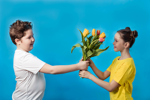 A teenage boy gives a bouquet of spring tulips to a pretty girl teenager on a blue background.