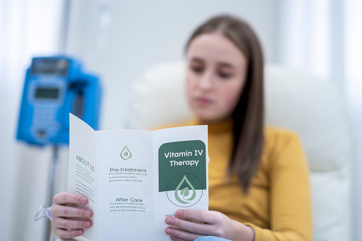 A female patient sits in an oversized high back chair as she casually reads a pamphlet on the vitamin treatment she is receiving.  She is dressed comfortably and has a neutral expression on her face as she receives her treatment intravenously.
