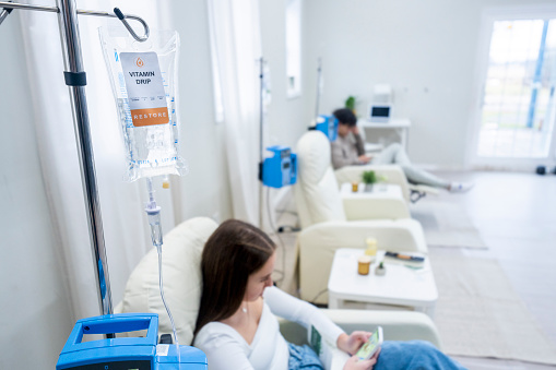 Two patients are seen in a Vitamin clinic as they sit in oversized high back chairs and receive their treatment intravenously.  They are each dressed comfortably as they occupy themselves with their phones and catch up on social media during the process.