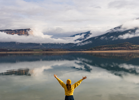 View from behind of unrecognizable excited woman with arms outstretched up wearing yellow sweater and wool hat looking at mountains reflected in lake