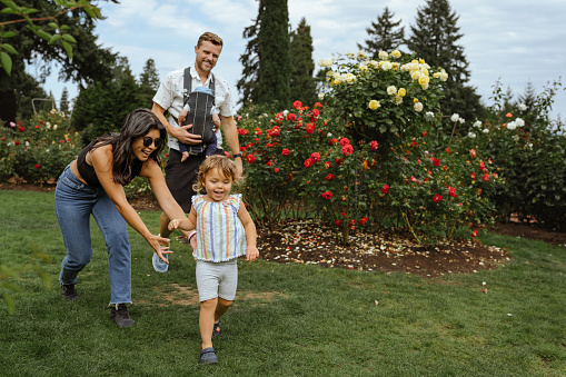 A woman of Latin American descent playfully chases her two year old toddler daughter through a flower garden while on a walk with her husband who is carrying their baby son in a baby carrier on his chest.