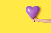 Woman hand holding a heart shape purple balloon on a yellow background