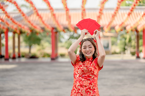 Happy Young lady wearing traditional cheongsam qipao costume holding ang pao, red envelopes in Chinese Buddhist temple. Celebrate Chinese lunar new year, festive season holiday