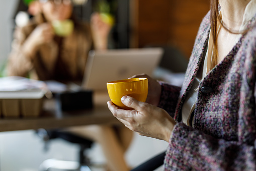 Selective focus shot of two young businesswomen sitting in a row, at their desks in a co-working office space, enjoying cups of coffee and casually chatting during a coffee break.