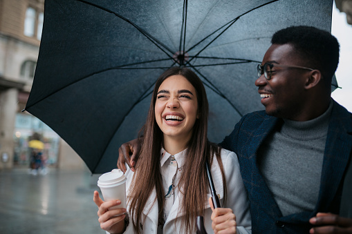 Young woman holding coffee and umbrella walking with male friends in the city on a rainy day. Interracial couple having fun walking around the city on a rainy day.