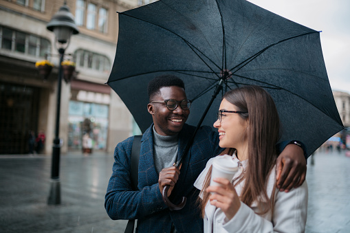 Loving young couple walking with umbrella and coffee in the city. Happy man and woman walking around the city on a rainy day.