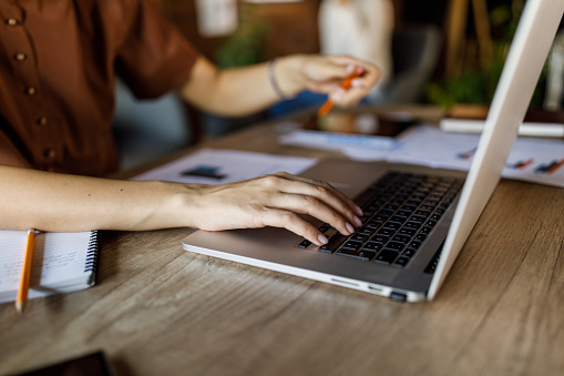 Close up shot of diligent young White woman sitting at her desk in a modern office space and typing data on laptop.