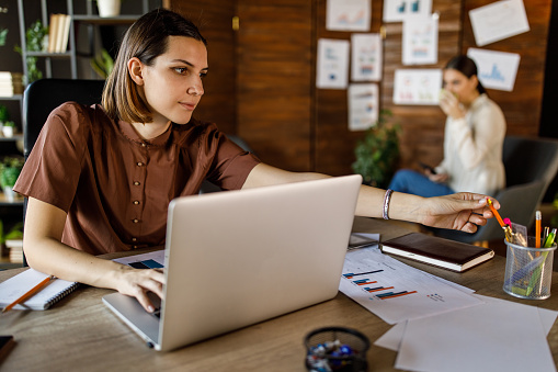 Portrait of diligent young White woman sitting at her desk in a modern office space and reaching for a pencil while typing data on laptop.