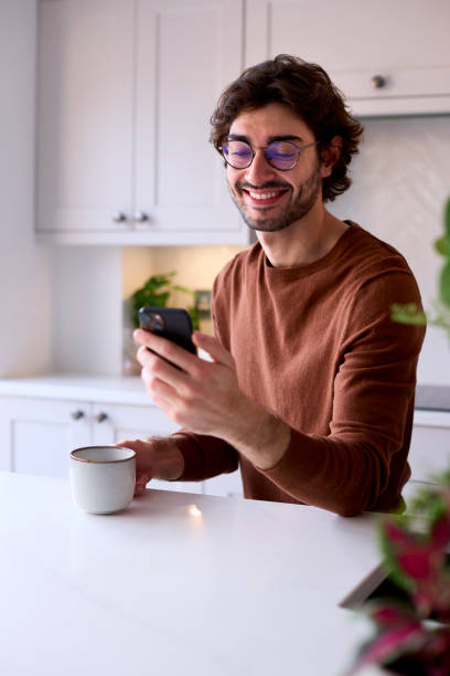 istock Young Man Relaxing At Home In Kitchen Holding Hot Drink Using Mobile Phone 1455773920