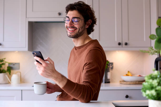 istock Young Man Relaxing At Home In Kitchen Holding Hot Drink Using Mobile Phone 1455773925