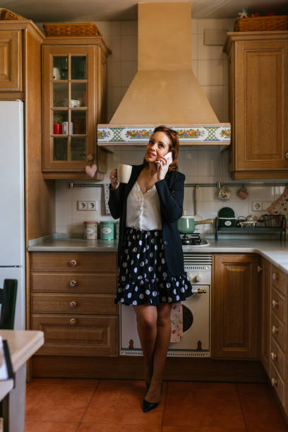istock Woman talking on mobile phone while drinking a cup of coffee in the kitchen at home. 1455773971