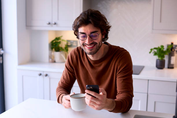 istock Young Man Relaxing At Home In Kitchen Holding Hot Drink Using Mobile Phone 1455773998