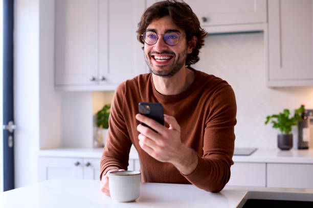 istock Young Man Relaxing At Home In Kitchen Holding Hot Drink Using Mobile Phone 1455774000