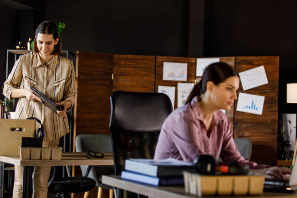 istock Young businesswoman working at her desk while her colleague is packing after getting fired 1455774087