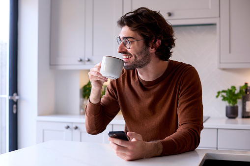 Young Man Relaxing At Home In Kitchen Holding Hot Drink Using Mobile Phone