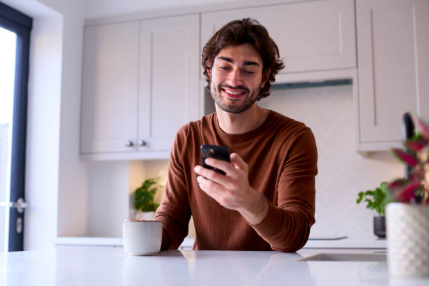 istock Young Man Relaxing At Home In Kitchen Holding Hot Drink Using Mobile Phone 1455774195