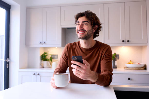 istock Young Man Relaxing At Home In Kitchen Holding Hot Drink Using Mobile Phone 1455774258