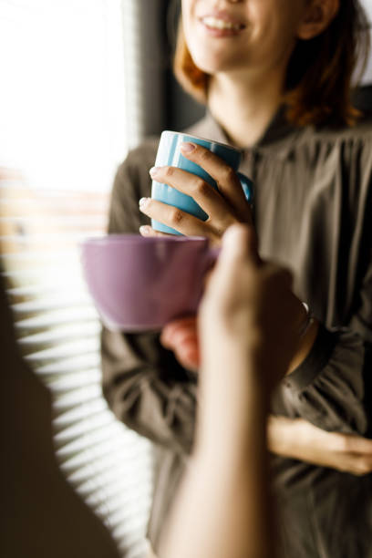Two young women standing by the window, having cups of coffee and chatting